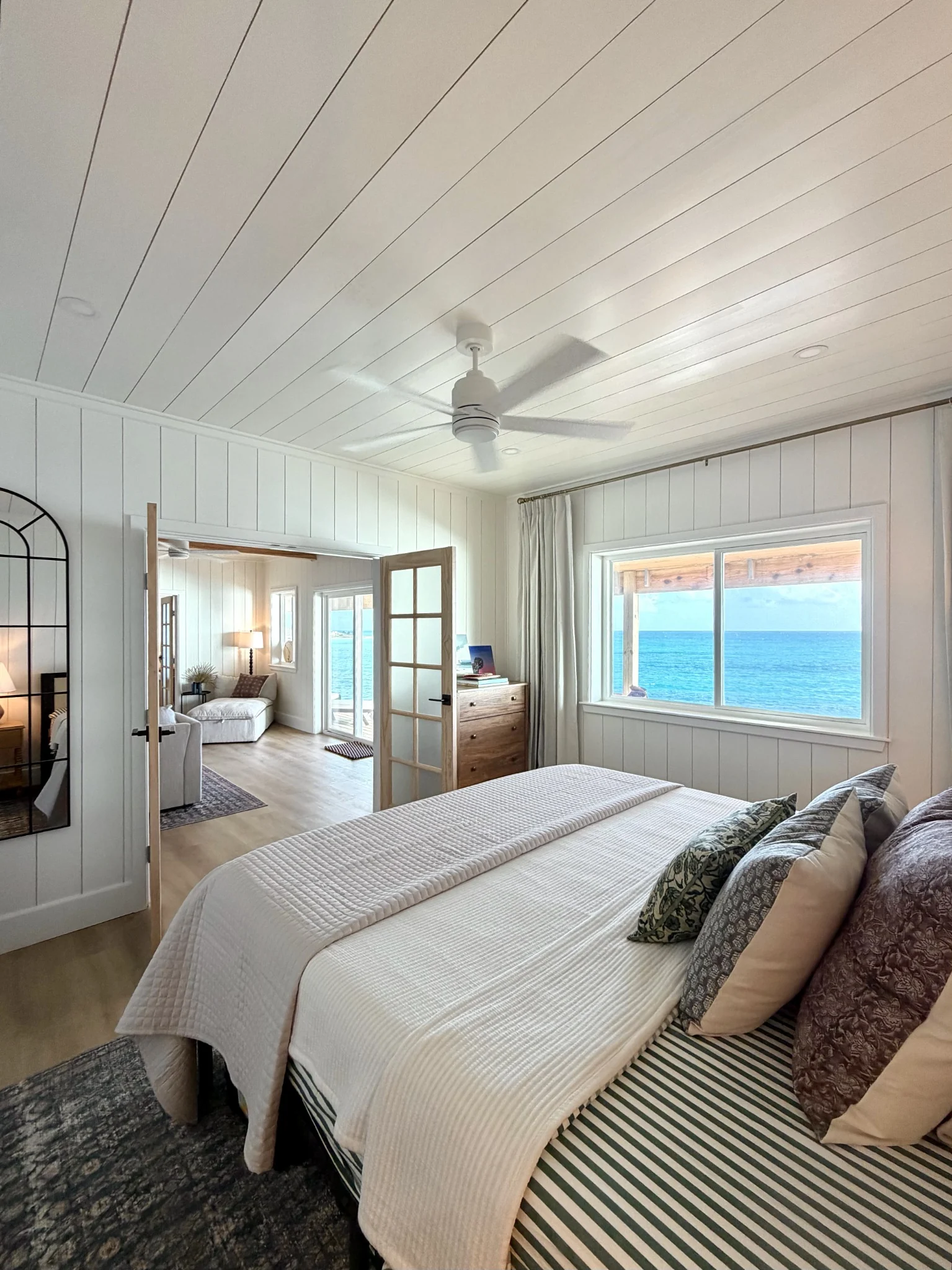 A white bed beneath a pale timber ceiling beside a window looking toward the ocean.