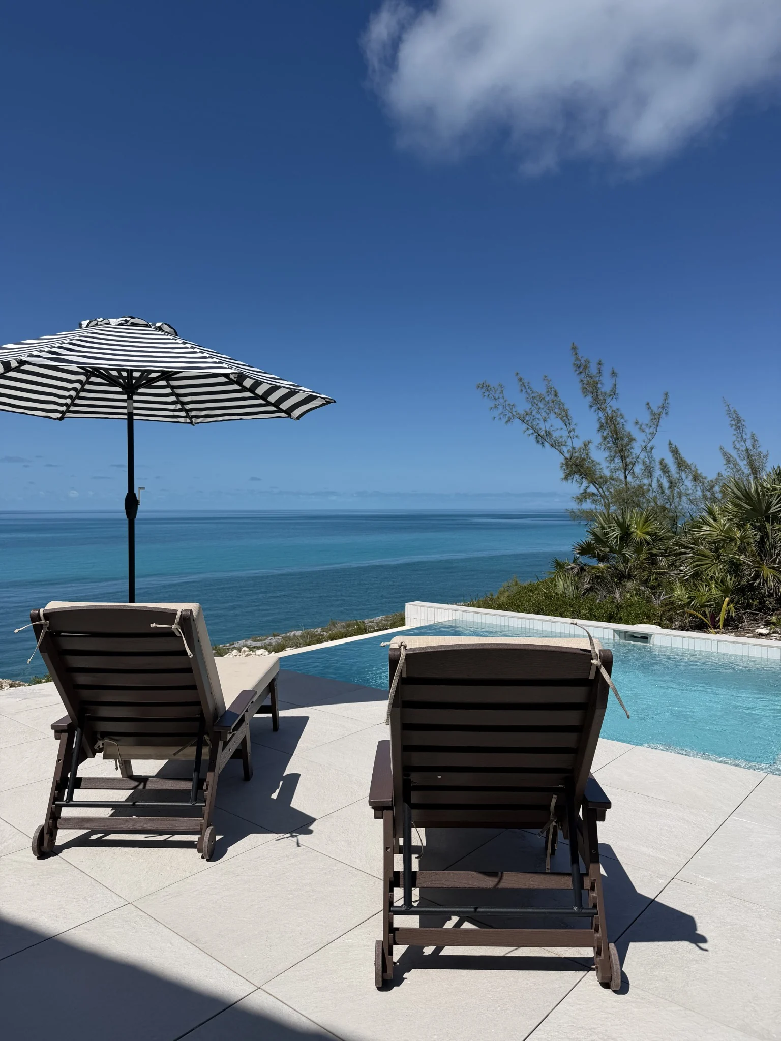 Two wooden loungers and a striped umbrella beside the CLIFFHOUSE pool overlooking the ocean.