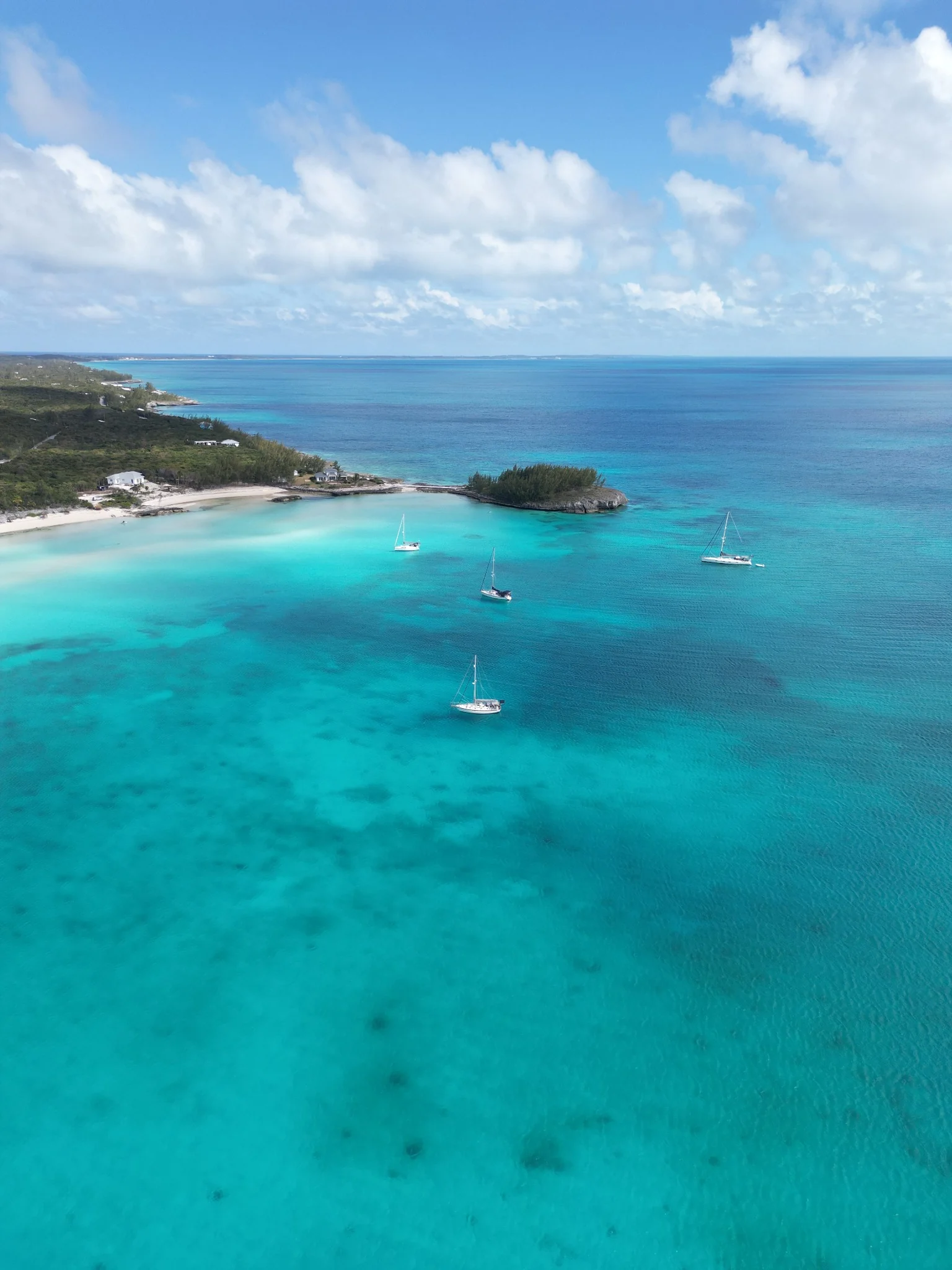 Sailboats anchored in the turquoise water of Rainbow Bay, Eleuthera, beside pale sand and a small green cay.